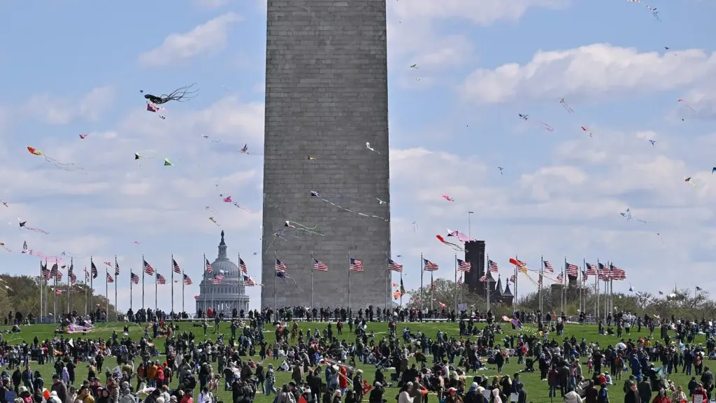 Crowds flying kites on the grounds of the Washington Monument with the Capitol dome in the distance
