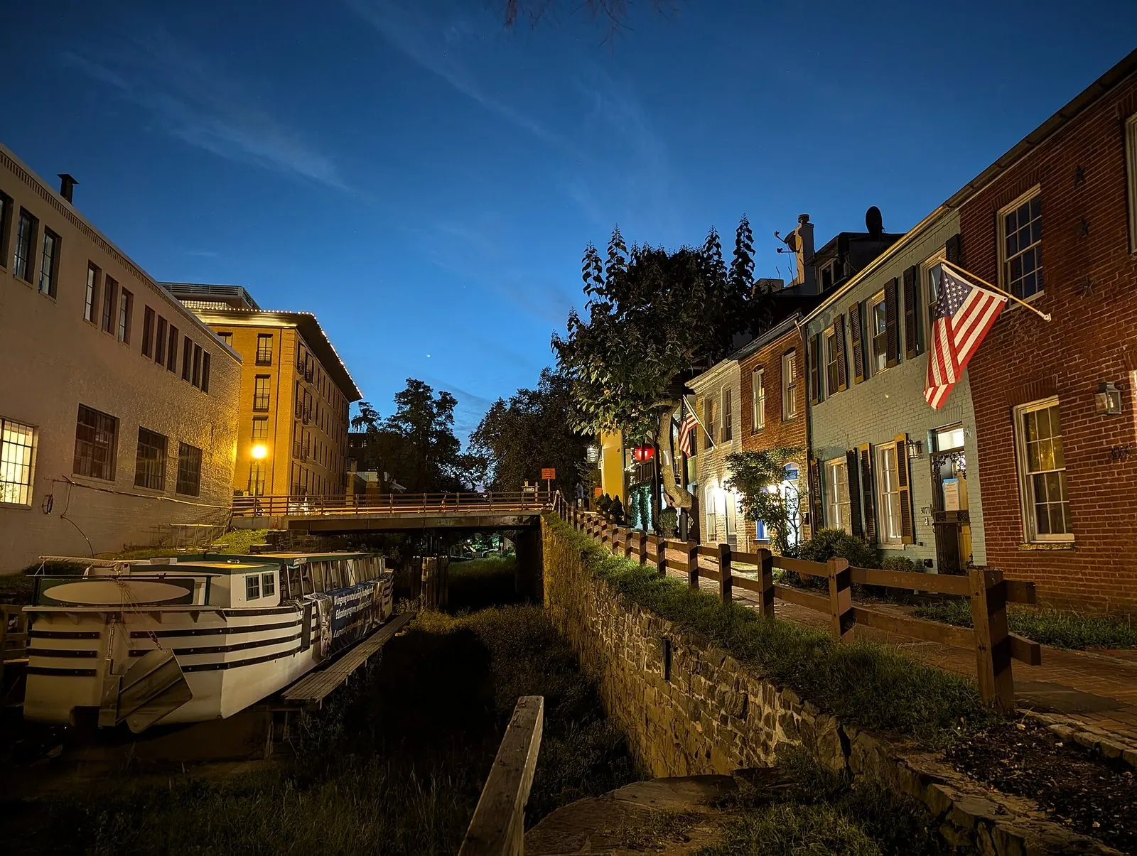 The C&O Canal in Georgetown DC at dusk, with a canal boat moored below brick rowhouses and a lit lamp