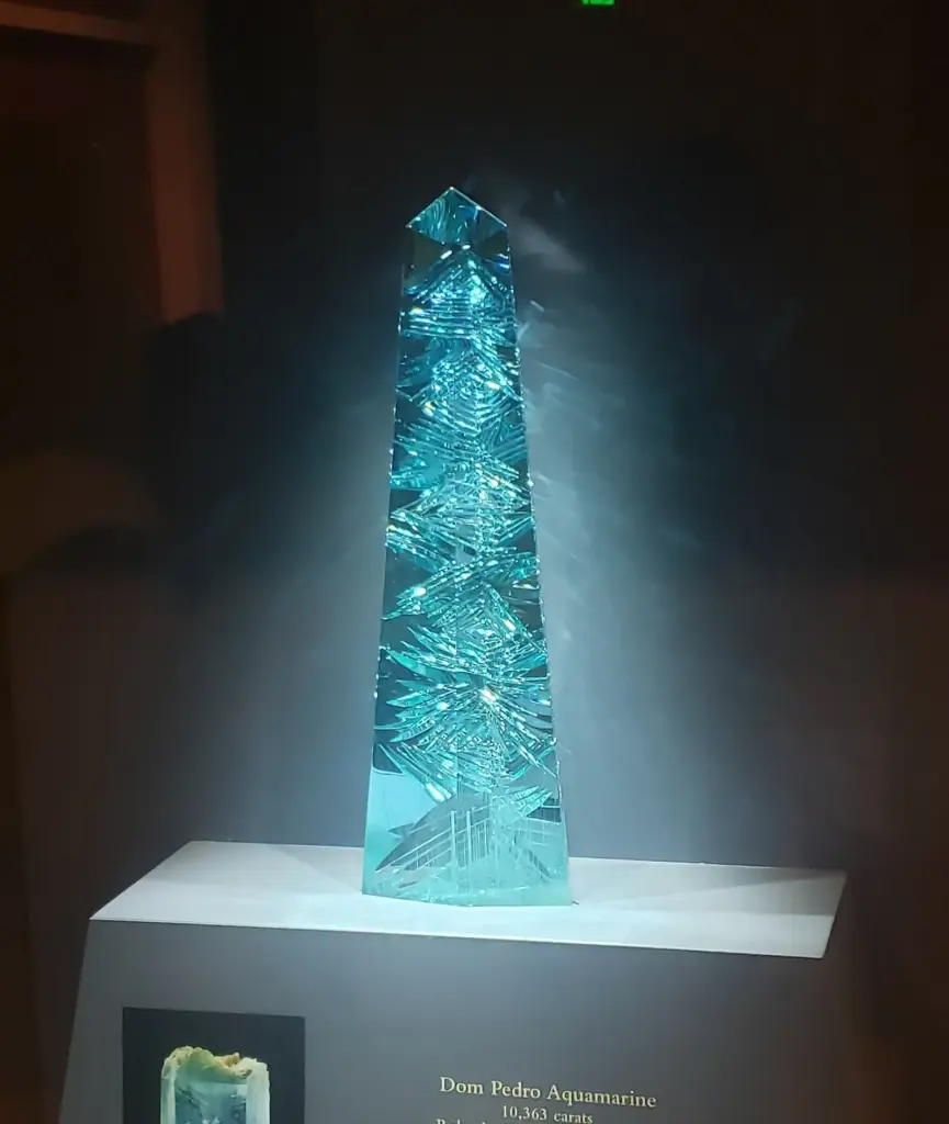 A tall pale blue aquamarine obelisk carved with fine facets, lit from above in a dark case