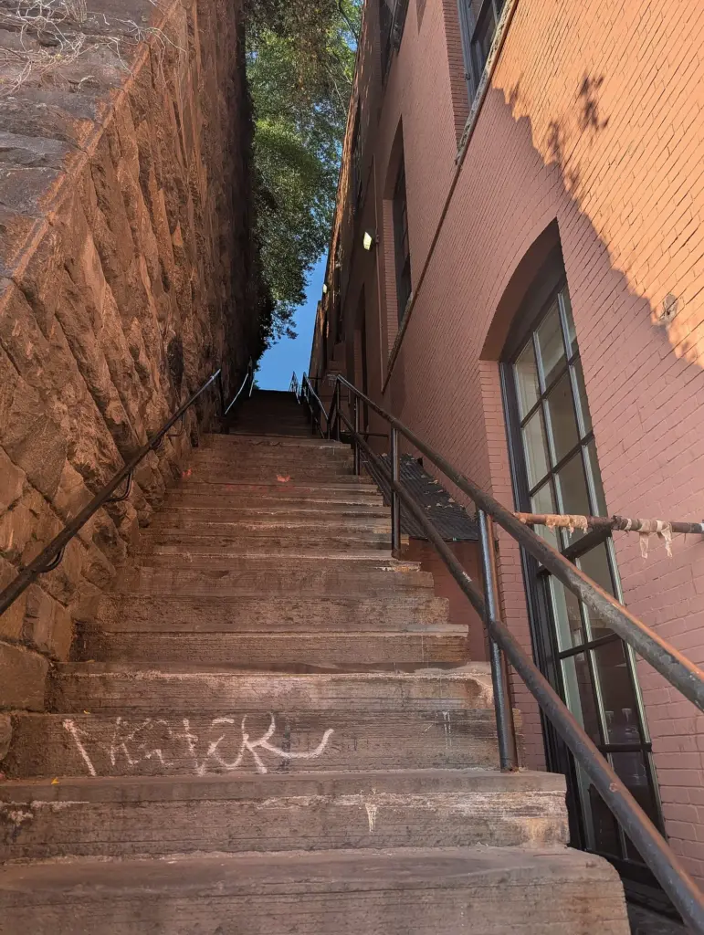 Steep stone staircase between a rough stone wall and a brick building, lit by afternoon sun