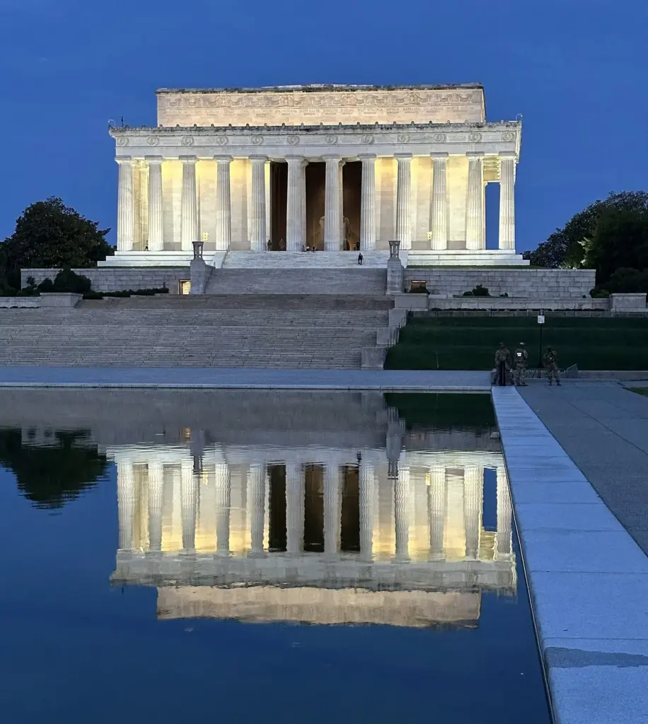 The white temple and its lit colonnade reflected in still blue water near sunrise