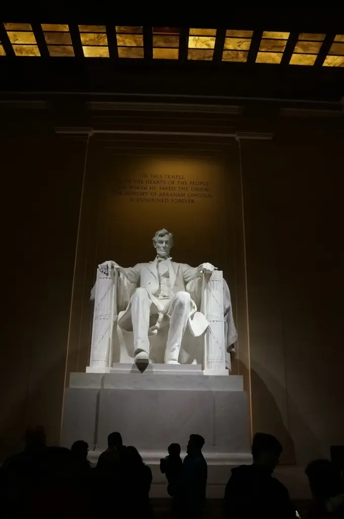 The lit statue of Abraham Lincoln inside the Lincoln Memorial at night, with visitors in silhouette
