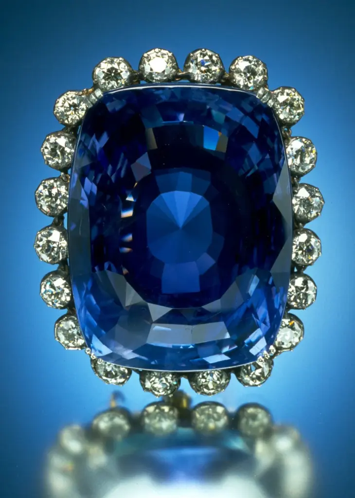 A large cushion-cut blue sapphire framed by a ring of round white diamonds against a blue background