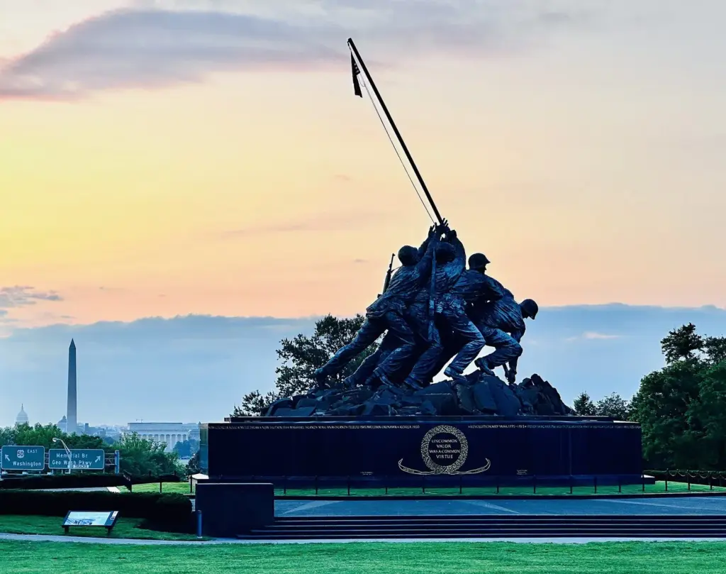 The bronze Marine Corps War Memorial statue at sunrise, with the Washington Monument and Capitol on the horizon