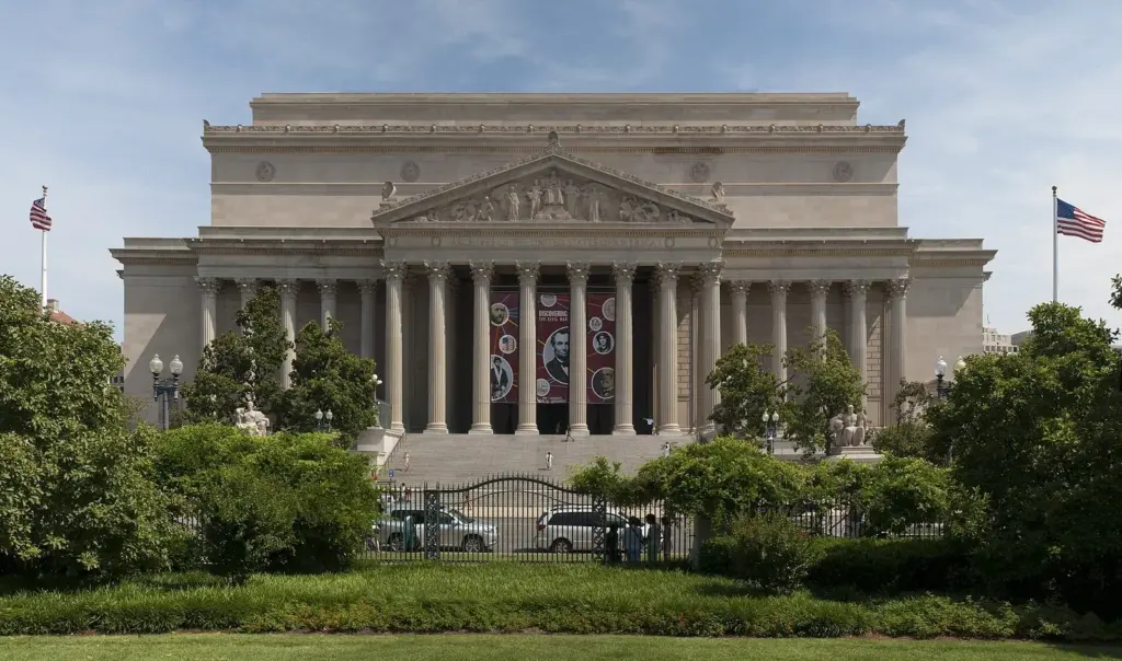 The classical columned facade of the National Archives Building behind trees and a fence