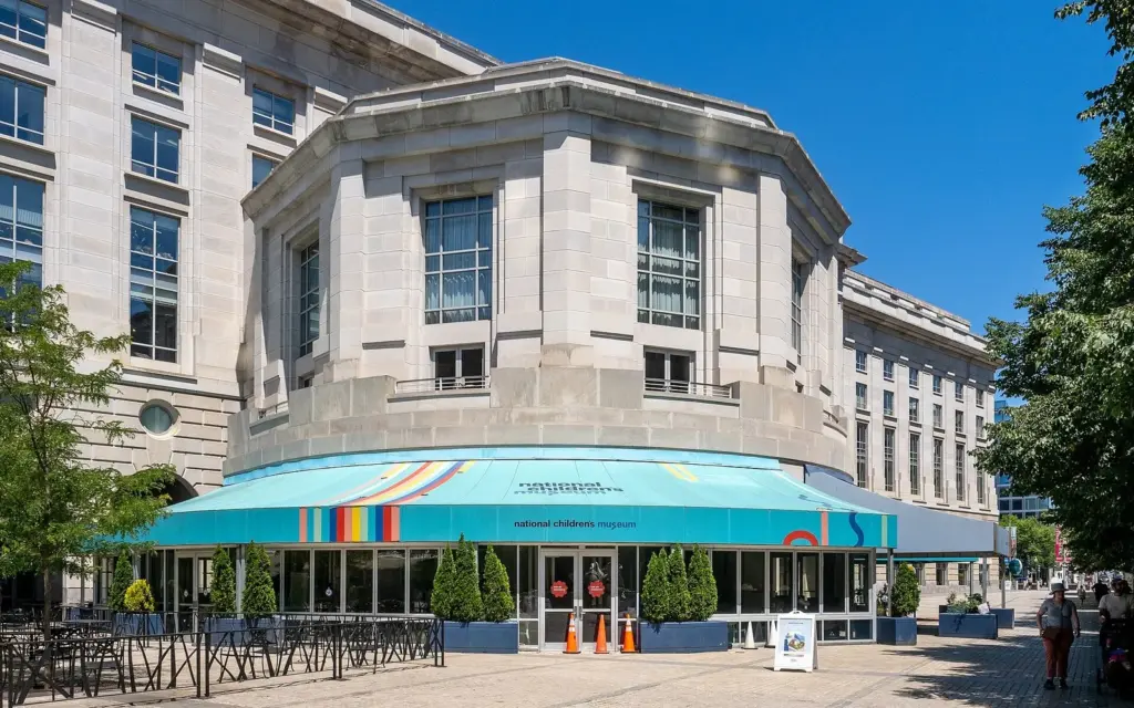 The curved glass entrance of the National Children's Museum under a colorful striped awning at the Reagan Building
