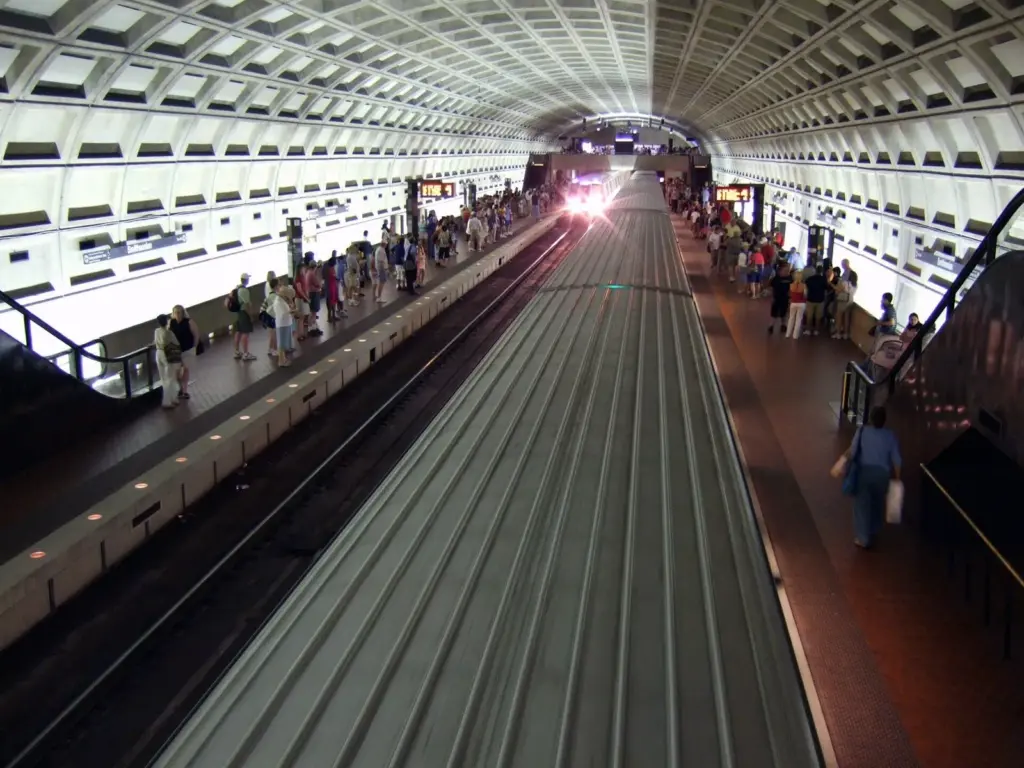 A train arriving under the coffered concrete vault of Smithsonian Metro station, with riders waiting on both platforms