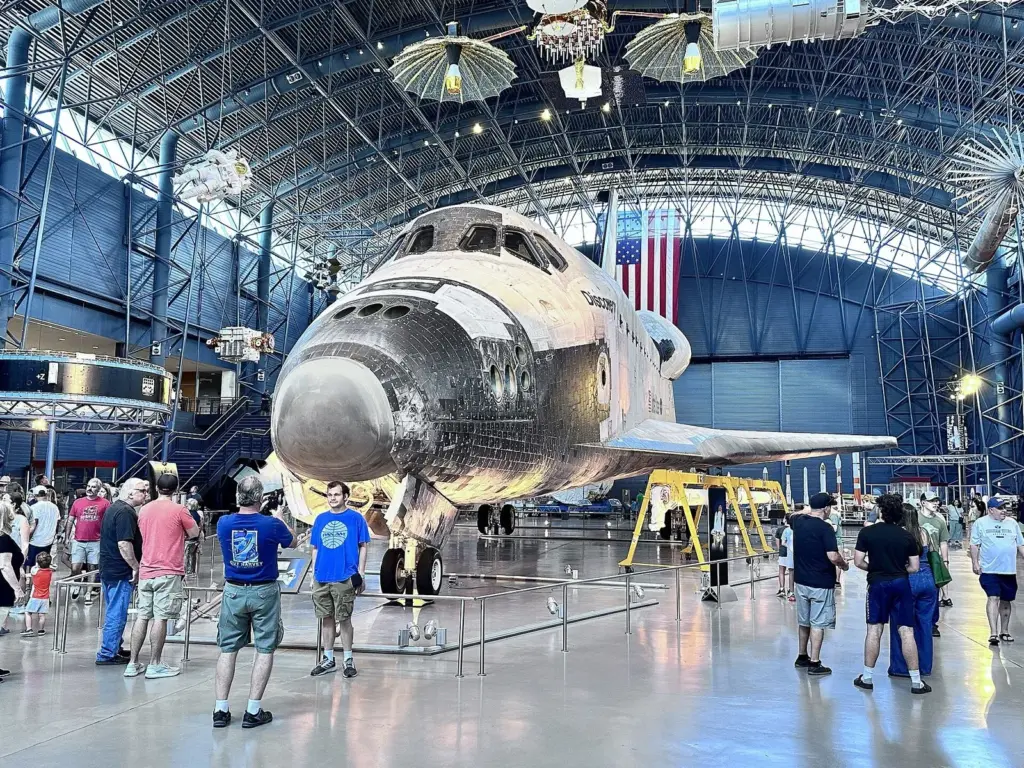 Space Shuttle Discovery on display in a vast hangar with visitors walking around its nose