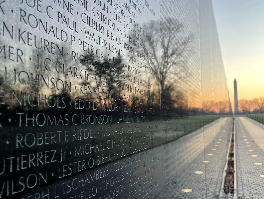 Rows of engraved names on the black granite Vietnam Veterans Memorial wall in low light, with the Washington Monument reflected